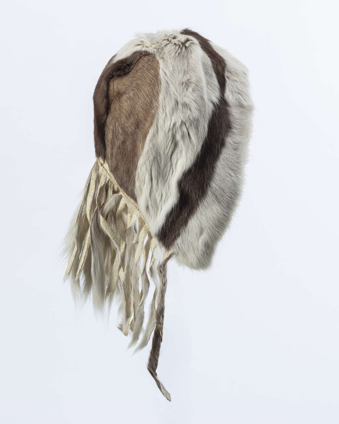 Capuchon en fourrure, vu latéralement, décoré de bandes verticales de fourrure blanche et brune. Des franges en cuir pendent au bas du capuchon.