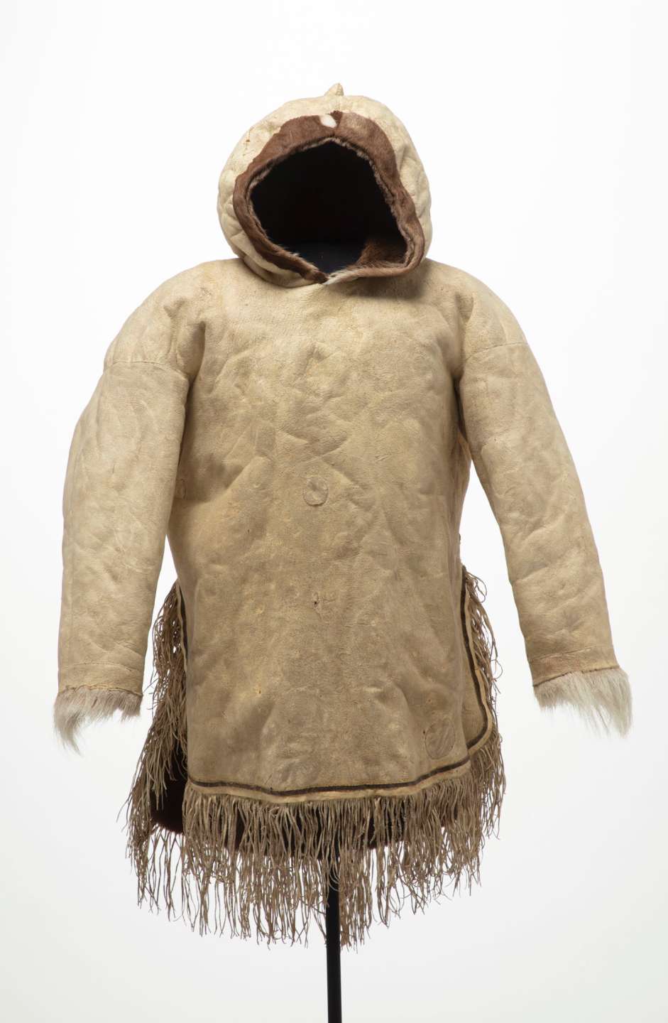 Veste en cuir beige vue de face, avec un capuchon pointu et relevé. La veste est fendue sur les côtés, et décorée de longues franges au bas et de fourrure blanche au niveau des poignets.