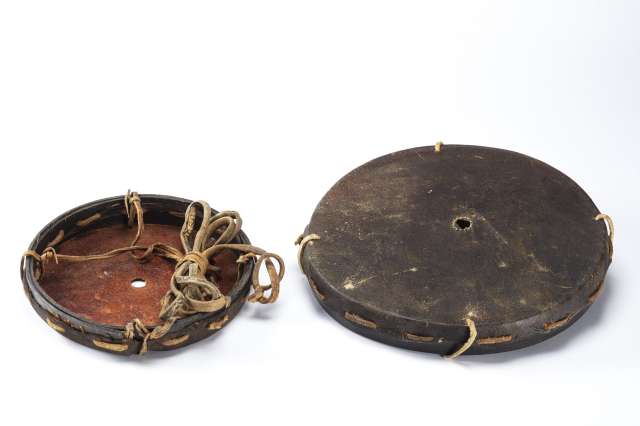 Deux objets circulaires à bords épais, en cuir brun foncé, tendu sur un cerclage. Tous les deux présentant un trou étroit au centre. L’un est renversé montrant l’intérieur rougeâtre et son cordage en cuir brun attaché en quatre points au cerclage.