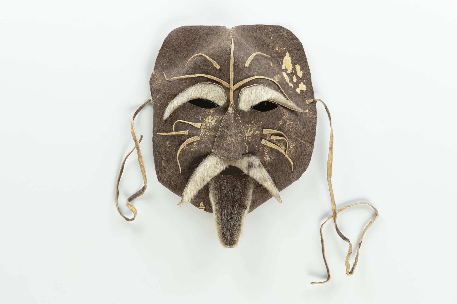 Masque en cuir brun, vu de face, avec des ouvertures pour les yeux et des sourcils, une moustache et une barbichette en fourrure. Le masque est orné de motifs de cuir en relief et est muni de cordons en cuir pour l’attacher.