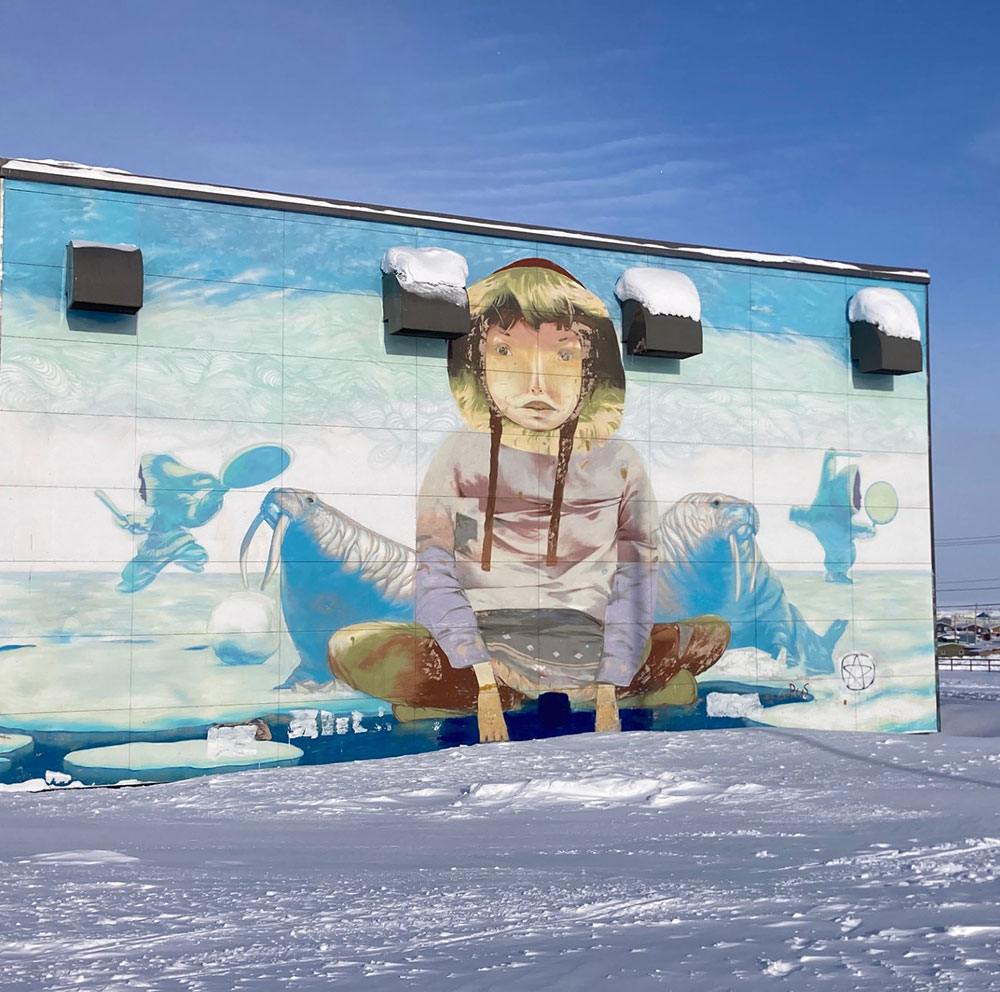 Cette photo illustre Igloolik en hiver : il y a de la neige au sol, une murale sur un bâtiment (à gauche) représentant un enfant assis, des morses et des danseurs. Deux personnes et plusieurs maisons apparaissent en arrière plan.