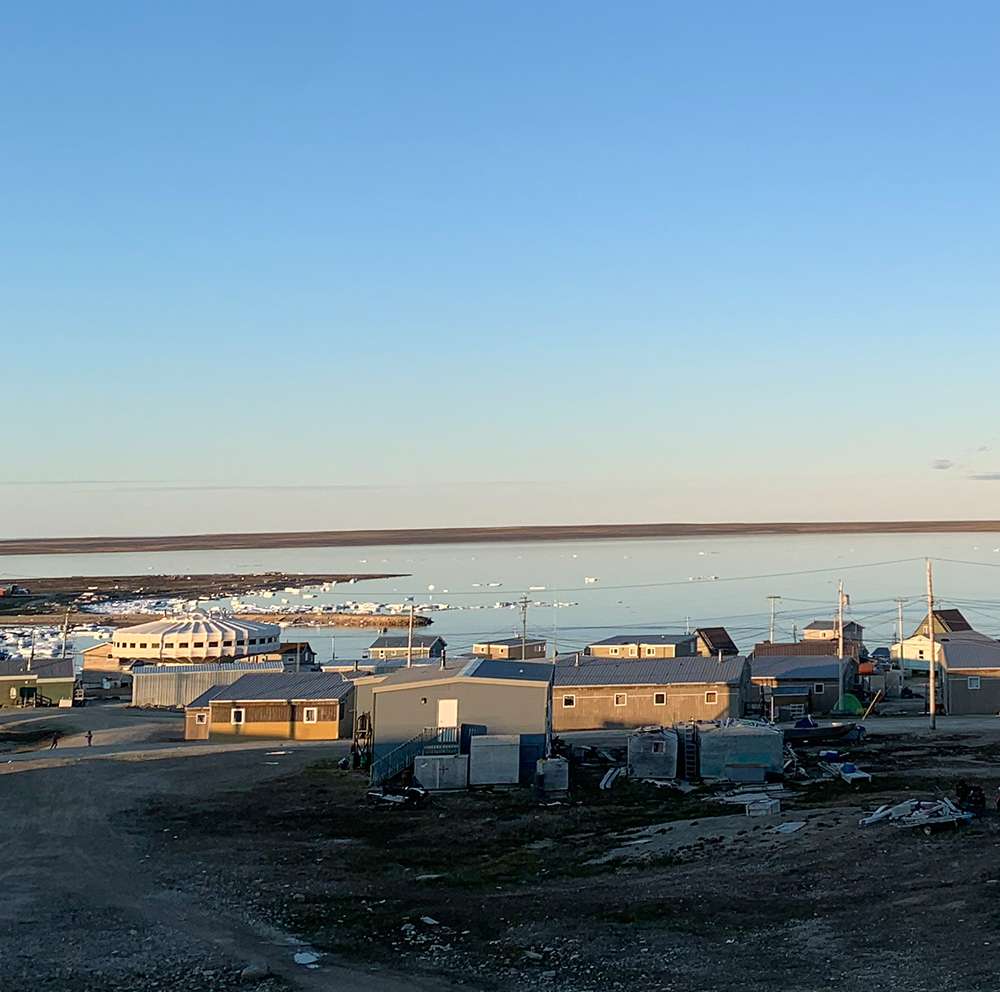 Cette photo propose une vue d'Igloolik en été: pas de neige au sol, plusieurs maisons toutes situées au bord de l'eau, un bâtiment en forme de champignon au milieu de celles-ci, et quelques morceaux de glace éparses au bord du rivage.
